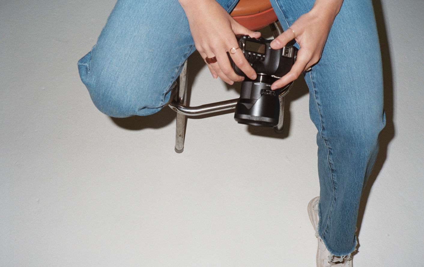Ashley seated on a metal stool in jeans and white sneakers, holding a digital camera across her lap.